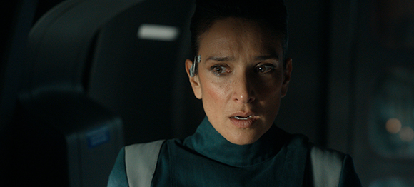A woman with short dark hair and a small device behind her ear looks ahead with a concerned expression.