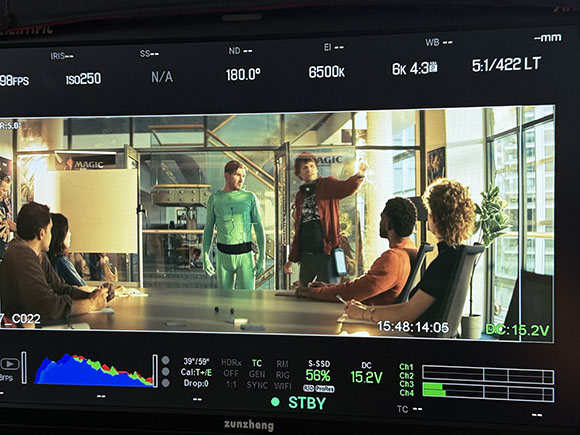 A camera monitor showing a film scene with actors in a glass-walled office and technical data overlays.