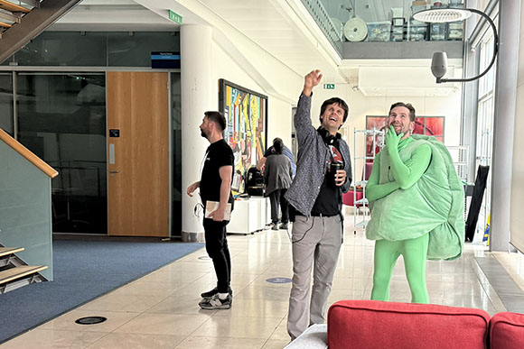A man in a green superhero costume laughs next to a man in a patterned shirt pointing upward in a bright lobby.
