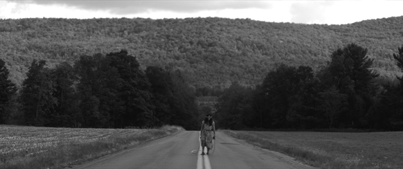 Black and white wide shot of a person walking down the center of a road toward a large, tree-covered hill.