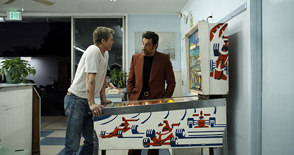 A man in a white t-shirt and jeans talks to a man in a brown blazer by an air hockey table.
