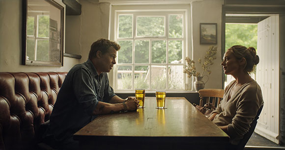 A man and woman sit across from each other at a wooden table with two glasses of beer in a dimly lit room.