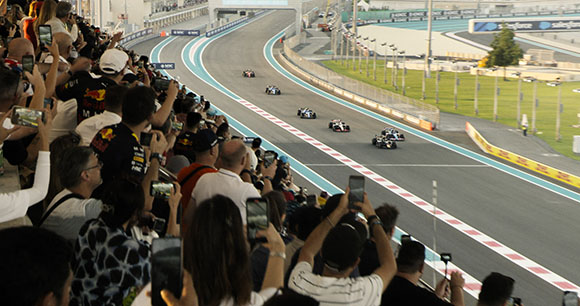 Race cars drive along a curved track past a crowd of spectators holding up smartphones.
