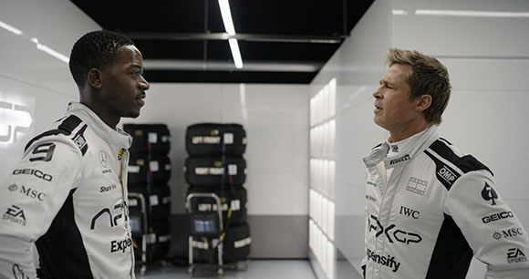 Two men wearing white and black racing suits stand facing each other in a brightly lit garage.