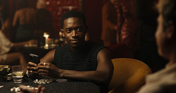A man with short hair and a sleeveless black top sits at a table in a dark, warm-toned lounge.