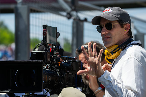 A man in a grey cap and sunglasses gestures with his hand while standing behind a large film camera.