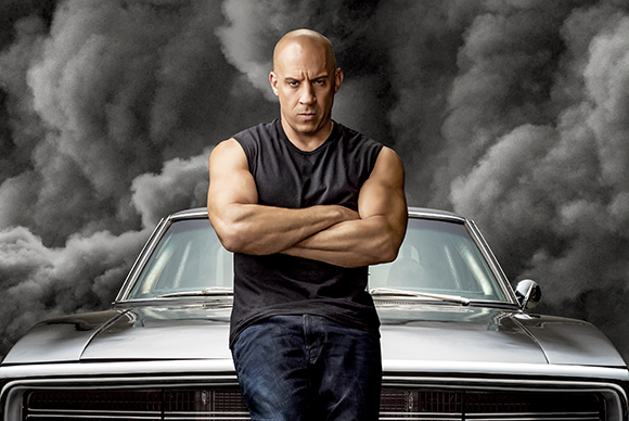 A muscular man in a black sleeveless shirt stands with arms crossed in front of a silver car and thick smoke.