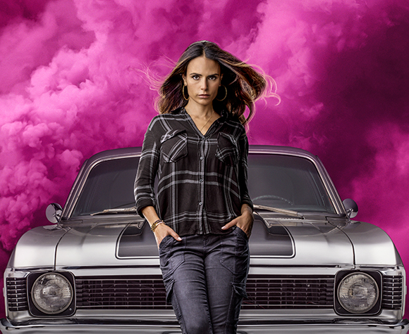 A woman in a plaid shirt and dark pants stands before a silver vintage car against pink smoke.