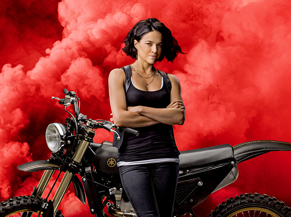 A woman in a black tank top stands with arms crossed in front of a motorcycle against red smoke.