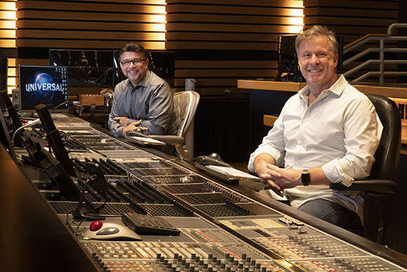 Two men smiling while seated behind a large professional audio mixing console in a studio.