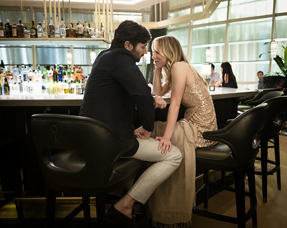 A man in a dark suit and a woman in a gold sequined dress sit on barstools talking.
