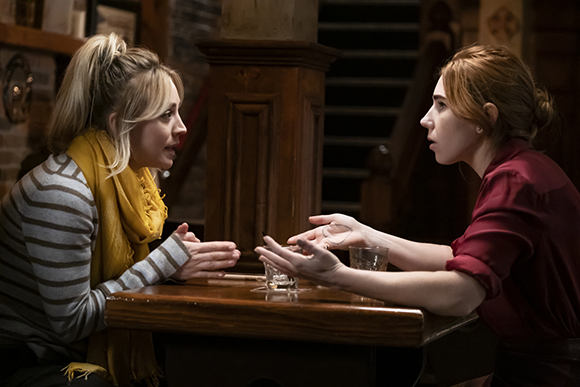 Two women sit facing each other at a dark wooden table in a dimly lit room, gesturing while talking.