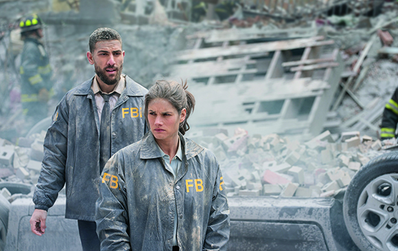 Two FBI agents in dusty jackets stand amidst rubble and debris at a disaster site.