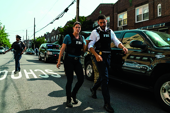 Two law enforcement officers in tactical vests walk past a black SUV on a city street.