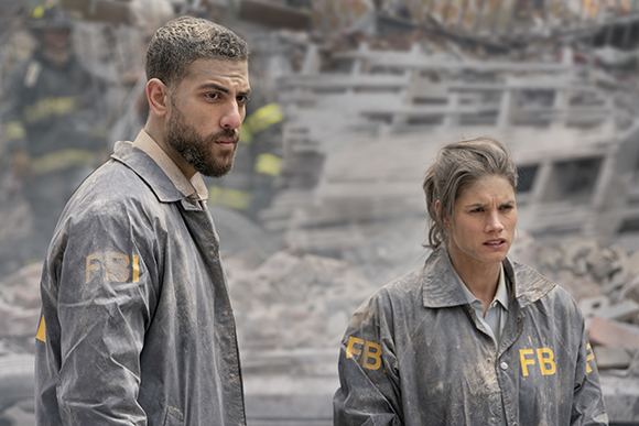 A man and woman in gray FBI jackets stand looking concerned in a field of debris.