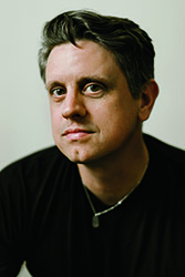 A man with dark hair and brown eyes wearing a black shirt and a silver necklace.