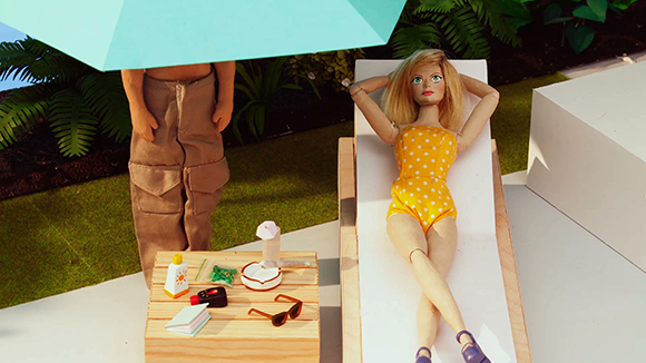 A blonde doll in a yellow polka dot swimsuit lies on a lounge chair next to a doll standing under a blue umbrella.