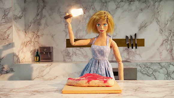 A blonde doll in a blue apron holds up a smartphone to photograph a piece of raw meat on a marble counter.