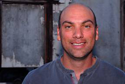A smiling bald man wearing a grey henley shirt against a weathered background.
