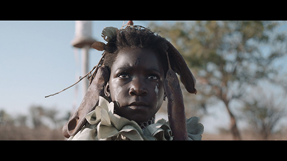 A young person with dark skin and facial markings looks forward, wearing leaf and wood adornments.