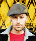 A man wearing a grey flat cap, a red shirt, and a dark jacket against a yellow patterned background.