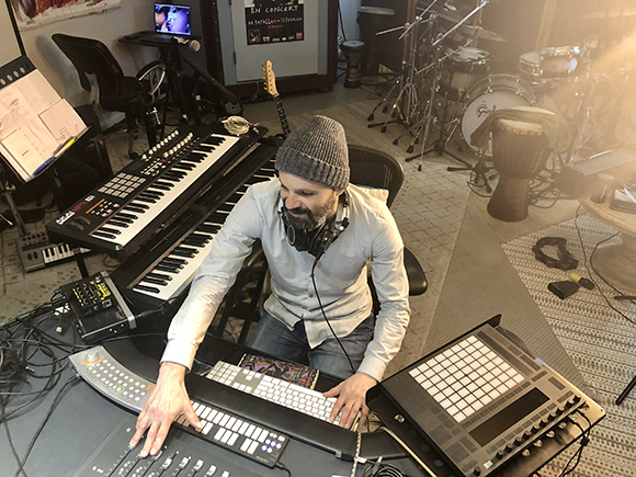 A man with a beard and grey beanie sits at a desk filled with musical keyboards and electronic controllers.
