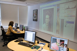 A person sits at a desk with computers while a large image of a portrait is projected onto the wall.