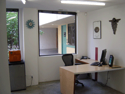 A small office with a light wood desk, black computer monitor, office chair, and a large interior window.