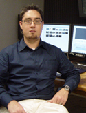 A man with glasses and a goatee wearing a dark blue button-down shirt sits before two computer monitors.