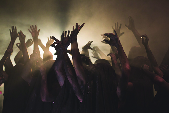 Silhouetted hands and arms raised toward a bright light in a dark, smoky crowd.