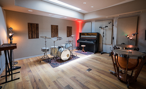 A music studio featuring a drum set, an upright piano, and large copper-colored timpani on a wood floor.