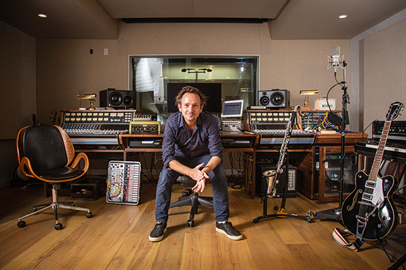 A man sits in a chair in a music studio filled with speakers, mixing consoles, and guitars.