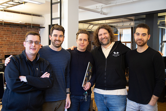 Five men standing side-by-side and smiling in a modern office with brick walls and large windows.