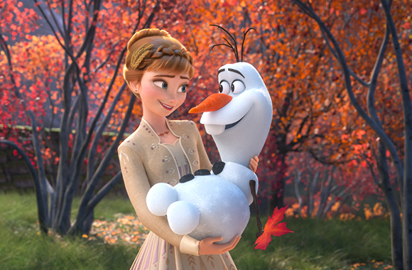 Anna smiles while holding Olaf the snowman in a forest with orange and red autumn leaves.