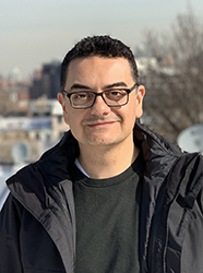 A man with short dark hair and glasses wearing a dark jacket and sweater outdoors.