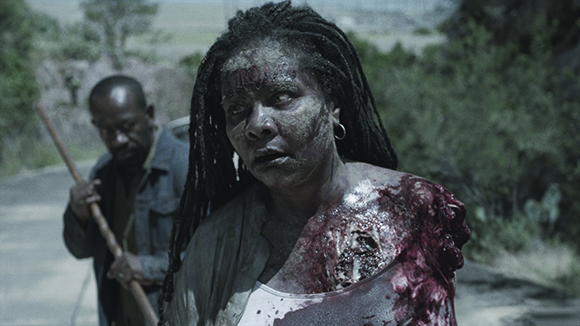 A woman with dreadlocks and blood on her chest looks distressed as a man stands behind her in a field.