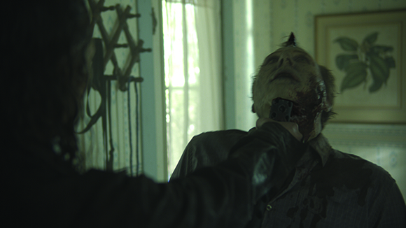 A person holds a dark device against the neck of a bloodied, pale-faced person in a dimly lit room.