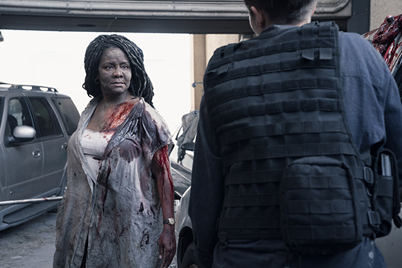 A blood-covered woman stares intensely at a person wearing a black tactical vest.