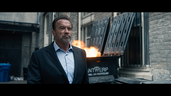 A middle-aged man in a dark suit and light blue shirt stands before a dumpster with flames rising from it.