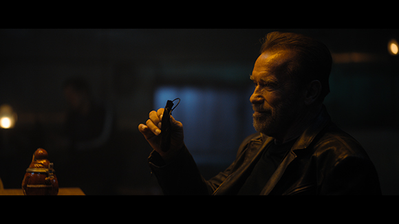 A man in a leather jacket holds a small black object in a dimly lit room.