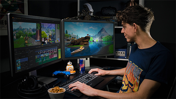 A young man uses a keyboard and mouse at a desk with two monitors displaying video game footage.