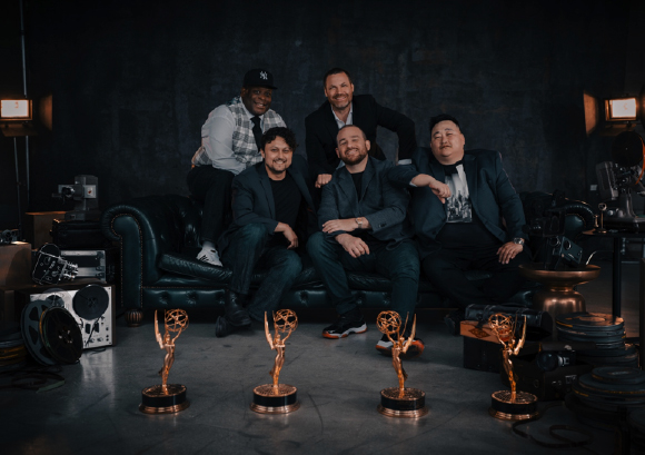 Five men smiling and posing behind four Emmy Awards in a dark studio.