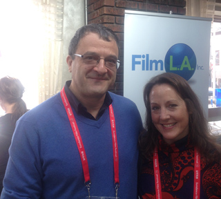 A man wearing glasses and a blue sweater stands next to a woman with long brown hair. They are both smiling and wearing lanya