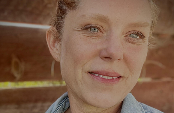 A close-up portrait of a woman with light eyes and reddish hair smiling against a wooden background.