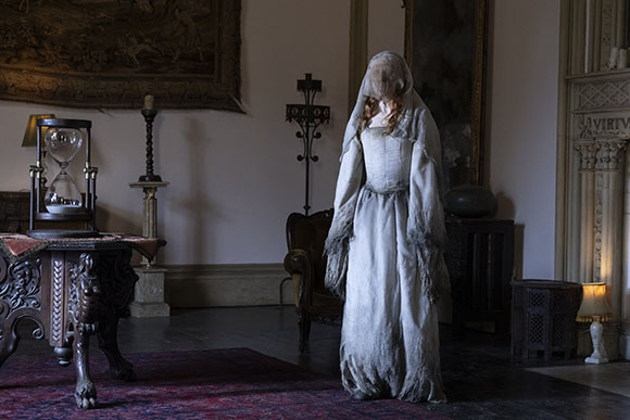 A person in a long, tattered white veil and dress stands in a dark, antique-filled room.