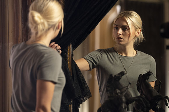 A blonde woman in a grey t-shirt looks at her reflection in a mirror framed by dark curtains.