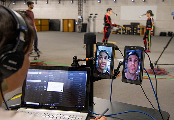 Person wearing headphones monitors two smartphones showing a man's face next to an open laptop in a studio.