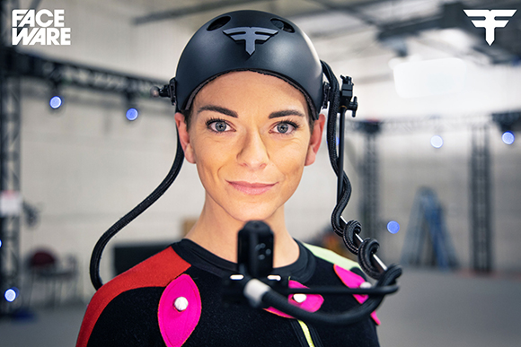 A woman wearing a black motion capture helmet and a black suit with pink accents in a studio.