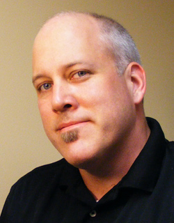 A man with light skin, blue eyes, and a small soul patch looking toward the camera.