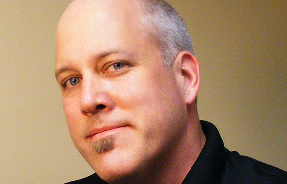 A close-up portrait of a bald man with blue eyes and a small goatee looking toward the camera.